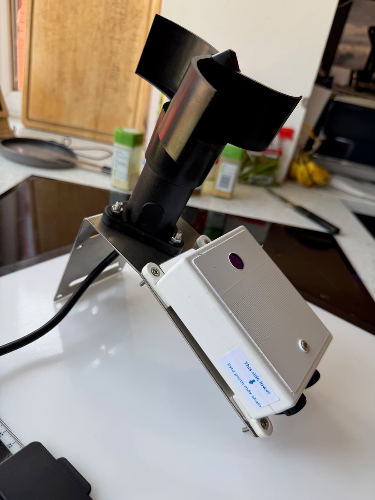 CloudWatcher and anemometer assembled on their metal bracket indoors, showing the angled sensor face and a “This side lower” label.