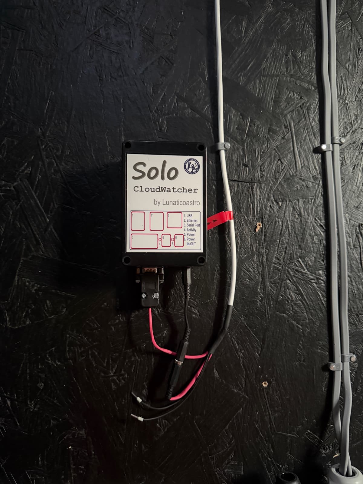 Solo CloudWatcher controller mounted on a black observatory wall, with its front connection legend and attached cables visible.